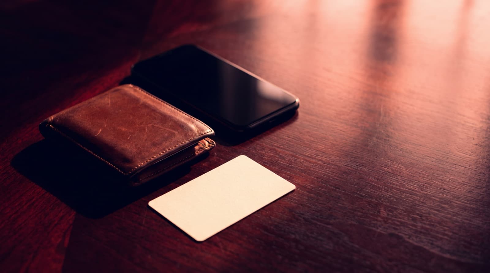 A worn leather wallet, a smartphone, and a blank card resting on a dark wood table in warm side light.