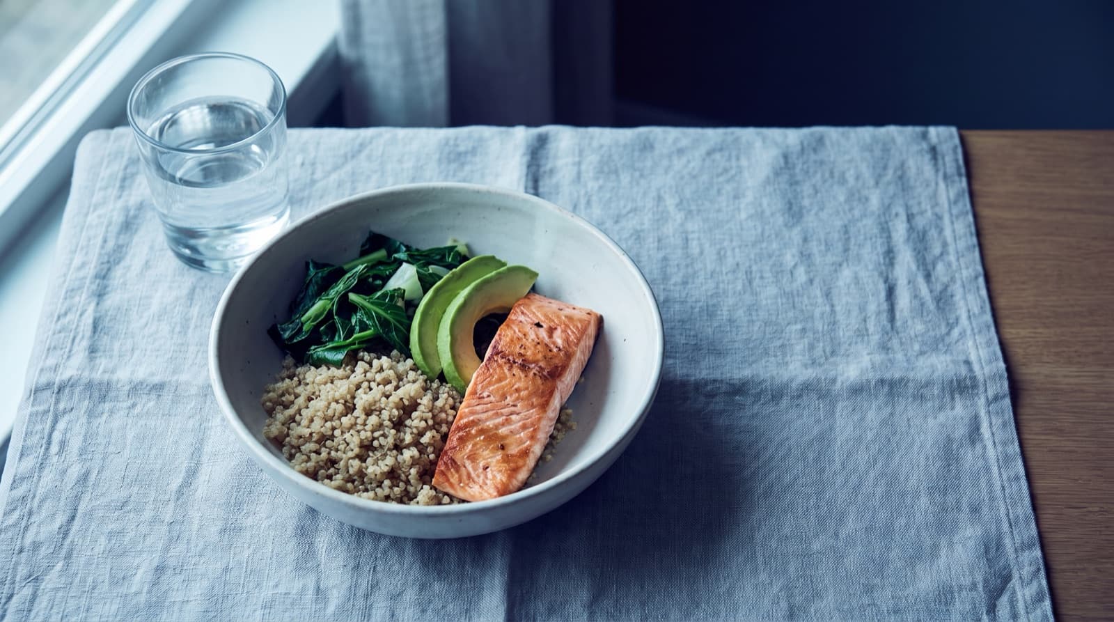 A ceramic bowl of salmon, quinoa, greens, and avocado beside a glass of water on a linen-covered table near a window.