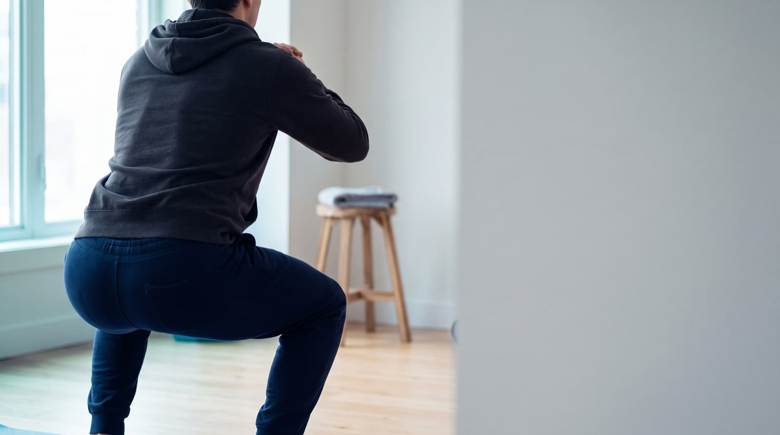 A person seen from behind, mid-squat in a grey hooded sweatshirt and navy joggers, in a bright empty room with a wooden stool holding a folded towel.