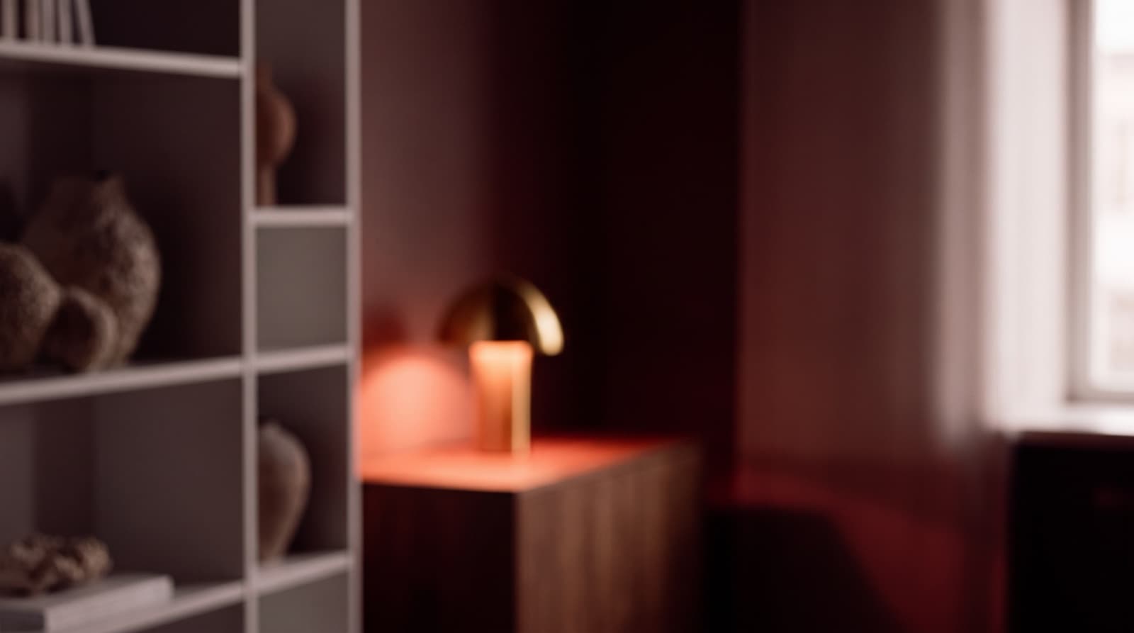 A warm, dimly lit room with a small lamp glowing on a dark wood console beside blurred ceramic vessels on a shelf.