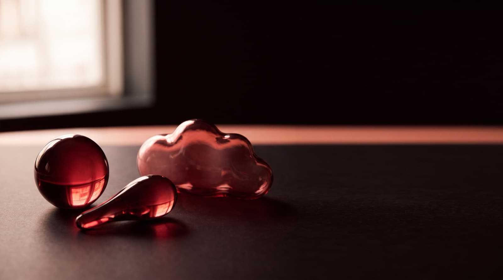 Three translucent red glass objects, a sphere, a rounded cloud shape, and a teardrop, resting on a dark table in window light.