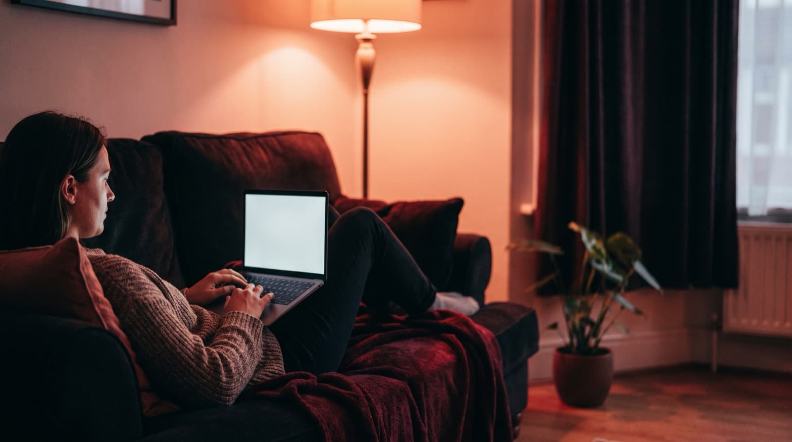 A person in a knit sweater reclines on a sofa in a warm lamplit living room, typing on a laptop.