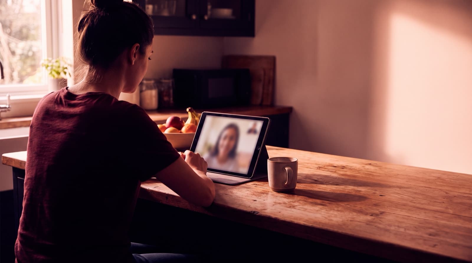 A person seen from behind at a kitchen counter, talking to someone on a video call on a propped-up tablet, with a mug beside them.