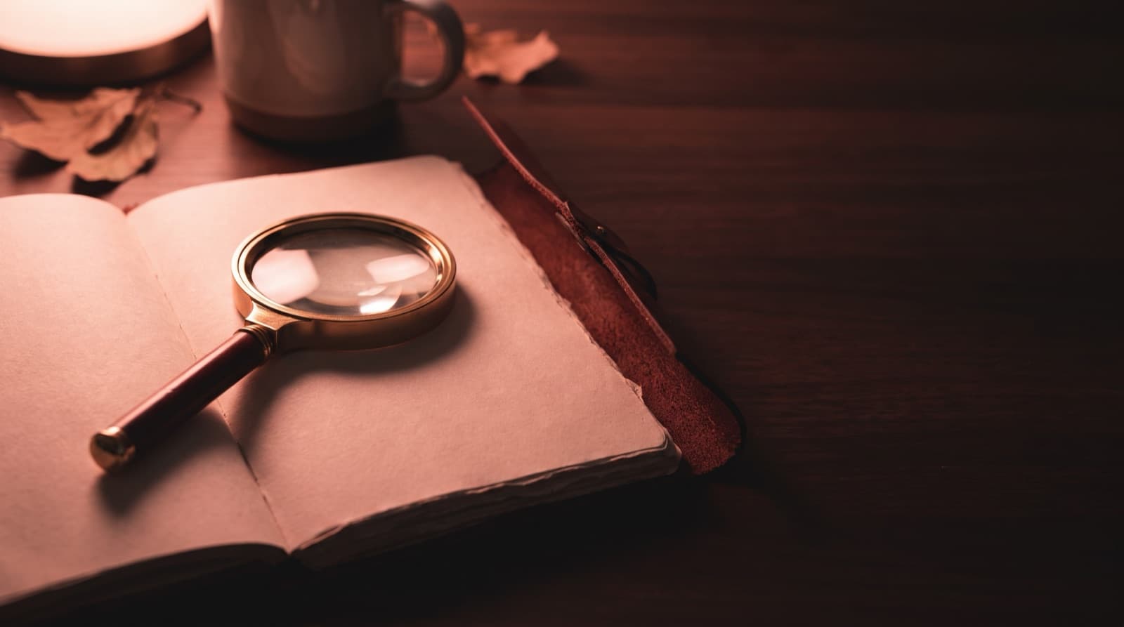 A brass magnifying glass resting on the blank pages of an open notebook on a dark wooden desk, next to a mug and a few dried leaves.