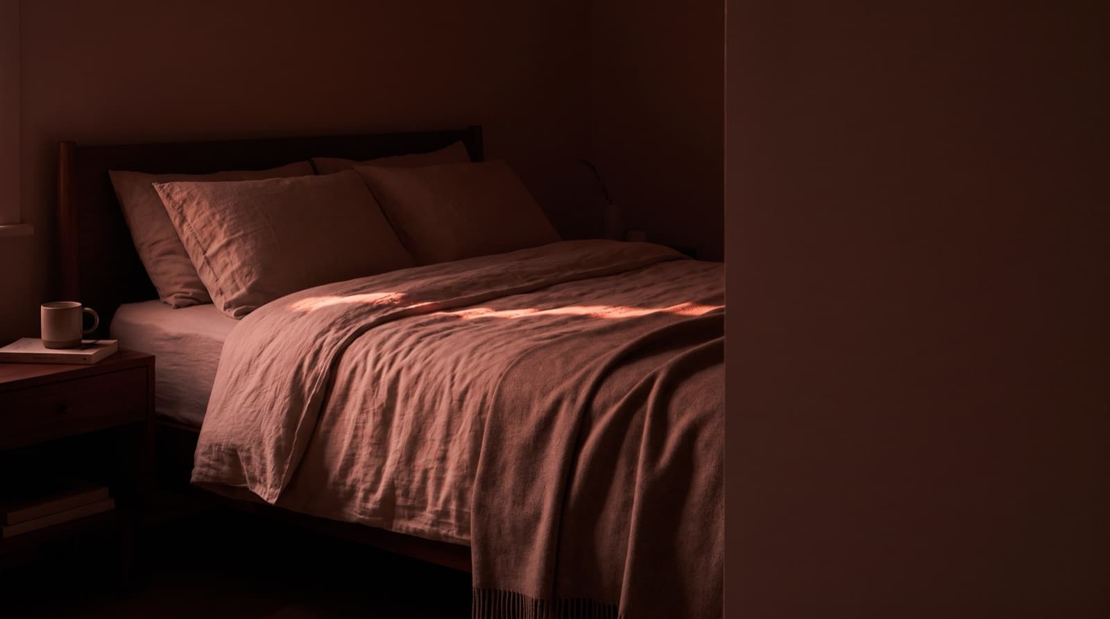A neatly made bed in a dim bedroom, with a stripe of warm light falling across the blanket and a mug and a book on the nightstand.