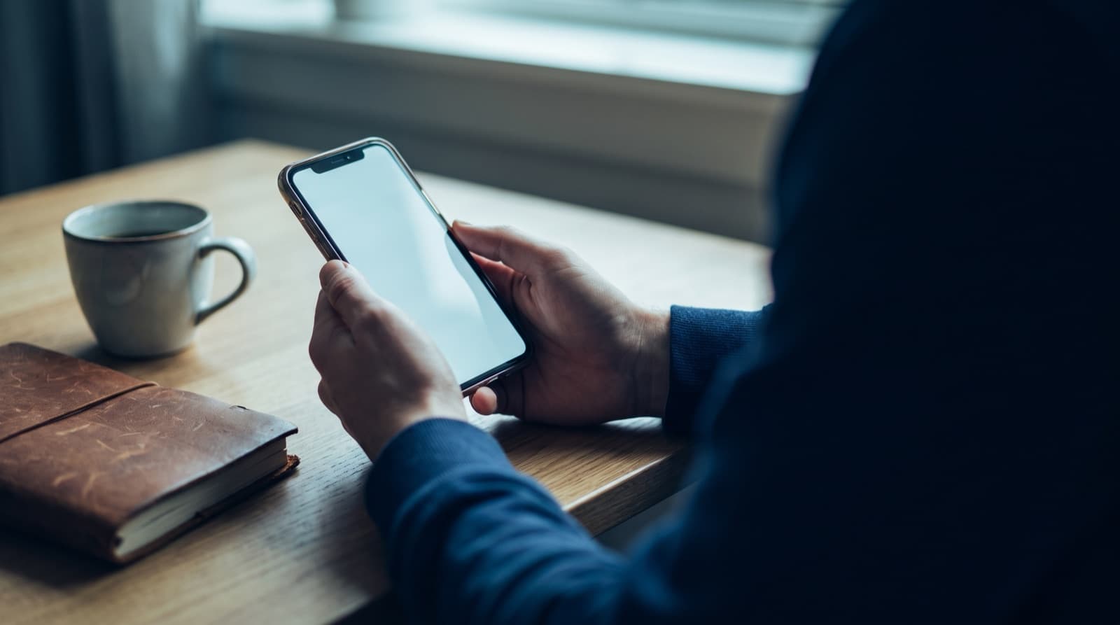 A person at a wooden table holds a smartphone with a blank glowing screen, a leather notebook and a cup of coffee resting nearby.