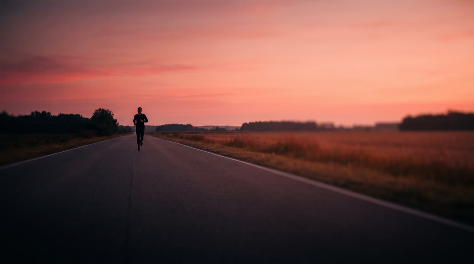 A lone runner on an empty rural road at dawn, small against a wide pink sky and open fields.